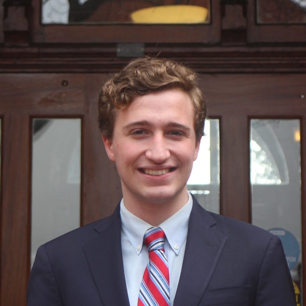 Ben Oestericher (SFS‘25) - Ben stands and smiles at the camera. He has short, slightly curly brown hair and is wearing a blue suit with a red striped tie. 