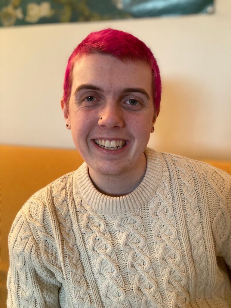 Elliot Lloyd (C‘24) - Elliot sits, facing the camera and smiling. He has short, pink hair and a nose piercing and is wearing a knit, cream colored sweater. 