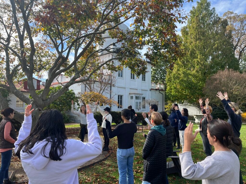 Students face the presenter and raise their arms as a presenter shares how to get in touch with nature. They are in a garden on a sunny fall day.