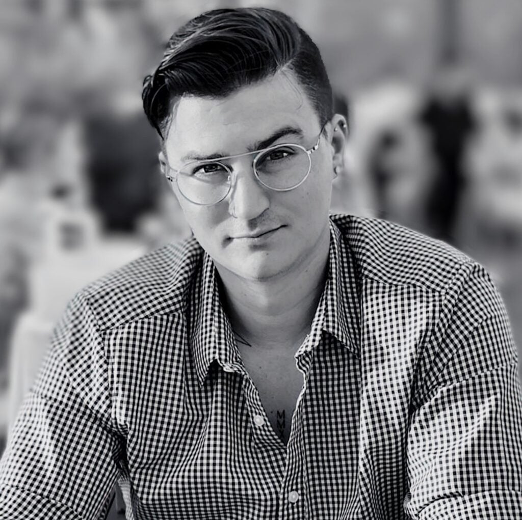 A black and white photo showing a person with light skin, short black hair, wire glasses, and a checkered button up shirt staring intently at the camera.